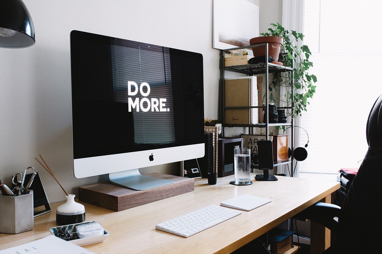 "Do More" is written in white capital letters on a black background on the screen of a MacBook computer. It's on a clean, neatly organised desk with a fresh light coming in from the right, outside of the shot. The bit of the room we can see feels modern and clean.