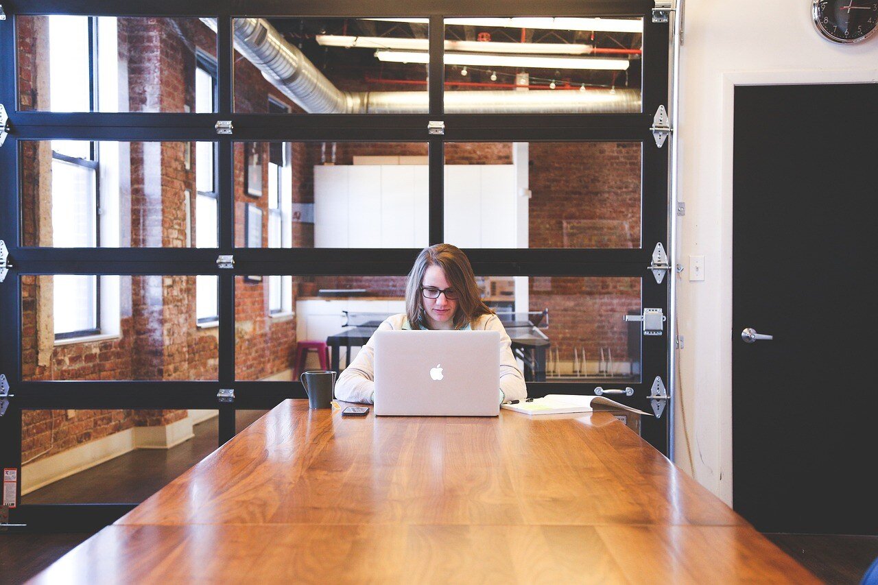 Brunette woman at a large wooden desk, looking at a macbook laptop, choosing her digital asset management software.